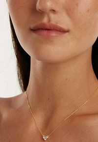 Collana d'oro con pendente a forma di cuore, caratterizzata da tre gemme trasparenti. Catena liscia, design delicato, indossata vicino alla clavicola.