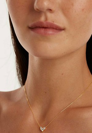 Gouden ketting met een hartvormige hanger, met drie heldere edelstenen. Soepele ketting, delicate uitstraling, gedragen nabij het sleutelbeen.