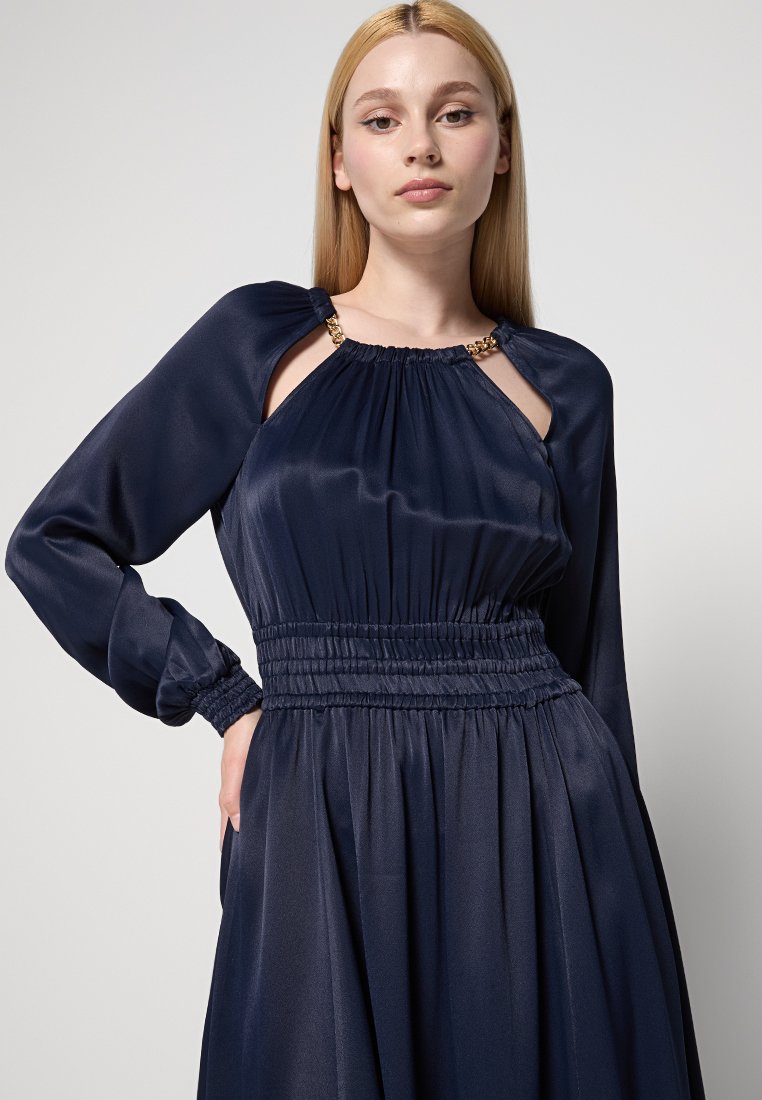 Vestito in satin blu navy con maniche lunghe, vita arricciata e dettagli cut-out sulle spalle; presenta accenti in metallo dorato.