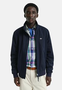 Giacca bomber blu navy con zip frontale, maniche e colletto a costine, sopra una camicia a quadri multicolore con bottoni, abbinata a pantaloni beige chiaro.