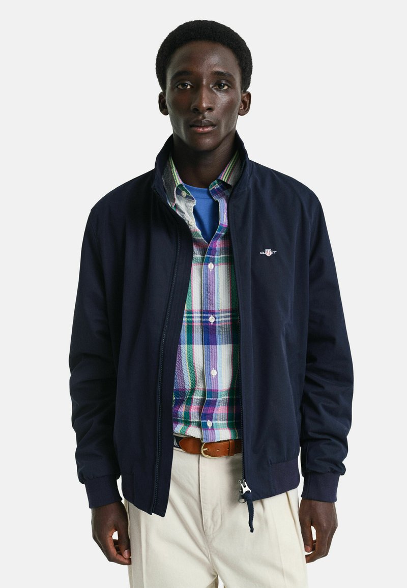 Giacca bomber blu navy con zip frontale, maniche e colletto a costine, sopra una camicia a quadri multicolore con bottoni, abbinata a pantaloni beige chiaro.