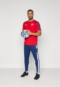 Maillot Adidas rouge avec des rayures bleues, pantalon marine avec des rayures blanches sur les côtés, tenant un ballon de soccer bleu et blanc, chaussures de sport noires.