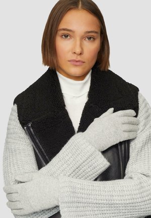 Vrouw met kort bruin haar, gekleed in grijze gebreide handschoenen, een grijze trui, een witte coltrui en een zwart schapenvacht kraagvest, met gekruiste armen.