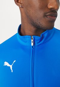Blauwe zip-up jas van gladde stof. Heeft een hoge kraag en een wit Puma-logo aan de linkerkant. Zilveren rits-hardware zichtbaar.