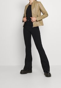 Giacca in pelle color cuoio con colletto rialzato e cerniere laterali, abbinata a pantaloni a vita alta neri con fondo svasato e scarpe platform nere.