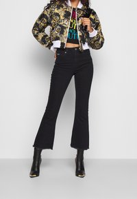 Svarta denimflaredjeans i kombination med en kort bomberjacka med ett guld- och svart blommönster och en färgglad grafisk t-shirt under.