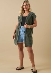 Ljusgrön, texturerad kofta med korta ärmar, öppen framsida, bärs över en svart linne och ljusa jeansshorts. Enkla sandaler och guldarmband.