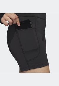 Ręka wkłada czarny smartfon do bocznej kieszeni czarnych dopasowanych szortów sportowych na jasnym tle.