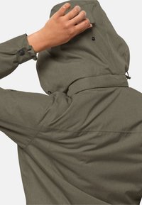 Person som justerar huvan på en olivgrön jacka med svarta tryckknappar och en dragsko mot en enfärgad bakgrund.