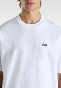 T-shirt en coton blanc avec un col rond et des manches courtes, orné d'un petit logo Vans noir sur la poitrine gauche.