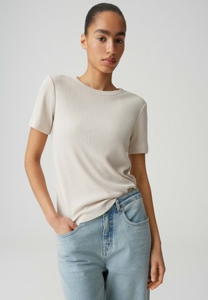 Junge Frau mit zurückgebundenem Haar, die ein geripptes beiges Kurzarmshirt und hellblaue Jeans trägt, stehend mit einer Hand in der Tasche.