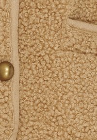 Beige tekstureret stof med en løkke-lignende, fuzzy overflade. Inkluderer en lille, rund messingknap og en sidelomme med en glat kant.