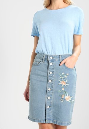 Woman wearing a light blue short-sleeve shirt tucked into a button-front denim skirt with floral embroidery on the right side.