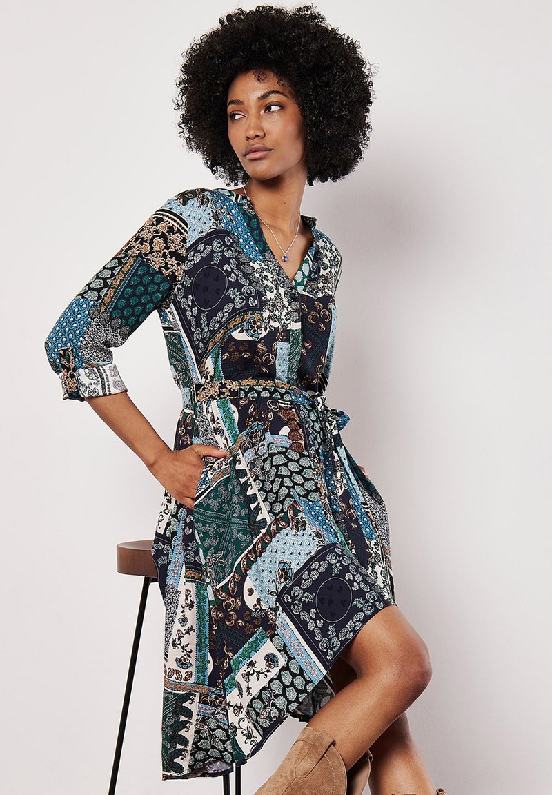 Dlouhý rukáv, kostkovaná šaty s kombinovanými vzory paisley a geometrickými, s páskem v pase, obsahující modrou, zelenou, černou a krémovou barvu.