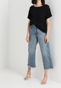Zwarte t-shirt met korte mouwen gecombineerd met lichtblauwe wijde jeans met rafelige zomen en beige strappy sandalen op een neutrale achtergrond.