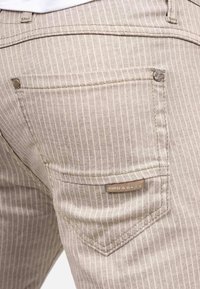 Pantalons rayés beiges avec une texture unique, des coutures visibles, une poche arrière avec une étiquette logo et un léger éclat.