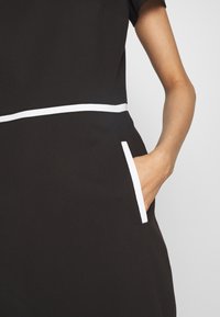 Gros plan sur une personne portant une robe noire avec des bordures blanches à la taille et à la poche, la main reposant à l'intérieur de la poche.