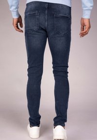 Person wearing dark blue slim-fit jeans and white sneakers, standing with hands relaxed at sides against plain background.