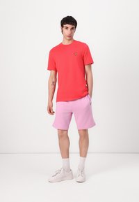 Röd bomullströja med en gul logotyp på bröstet, kombinerad med rosa bomullsshorts. Modellen bär vita sneakers och strumpor.