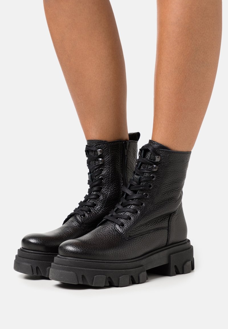 Bottes de combat en cuir noir texturé avec des semelles à crampons épaisses et des lacets, portées sur des jambes nues devant un fond clair uni.