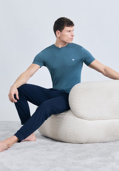 Άνδρας με μπλε-πρασινωτό μπλουζάκι και σκούρο μπλε παντελόνι κάθεται ξυπόλητος, στρίβοντας τον κορμό με τα χέρια τεντωμένα, ακουμπώντας σε κρεμ καναπέ δύο κομματιών με υφή.