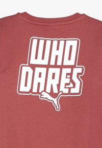 Camiseta marrón con texto en blanco en negrita "WHO DARES" y logo de Puma. Fabricada en tejido suave, con cuello redondo y mangas cortas.