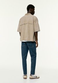 Chemise beige à manches courtes avec un empiècement au dos, associée à un jean en denim bleu. La tenue présente une coupe décontractée et des baskets contrastées.