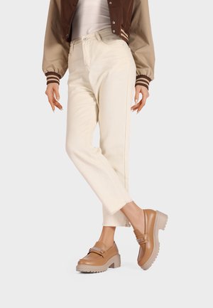 Jeans dritti color beige chiaro con orli sfrangiati, abbinati a mocassini con plateau marroni dotati di una fascia decorativa e suola testurizzata.