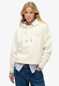 Sweat à capuche couleur crème avec une grande poche avant, capuche à cordon de serrage et logo Superdry. Porté par-dessus une chemise à rayures bleues et blanches, associé à un jean foncé.