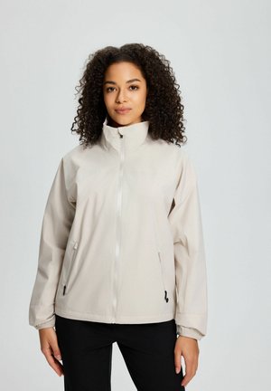 Frau mit lockigem Haar, die eine hellbeige Jacke mit Reißverschluss und Taschen trägt und vor einem schlichten hellgrauen Hintergrund steht.