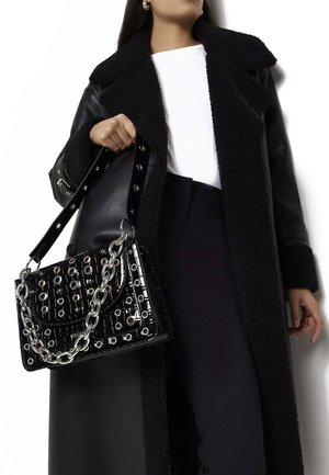 Sort håndtaske med blank overflade, sølv kæde rem og dekorativt hulmønster. Båret med en sort shearling frakke og en hvid top.