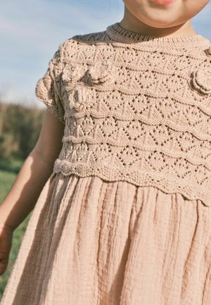 Stickad klänning med en texturerad beige överdel som har blommiga detaljer och korta ärmar, som fortsätter i en plisserad, ljusbeige kjol.