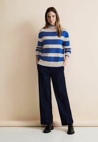 Pull à rayures bleu et blanc avec un col haut et un ourlet côtelé, associé à un pantalon navy à jambes larges et des chaussures noires.