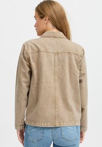 Lysbrun denimjakke med løstsiddende pasform, knaplukning, synlige sømme og lille krave, parret med blå jeans.