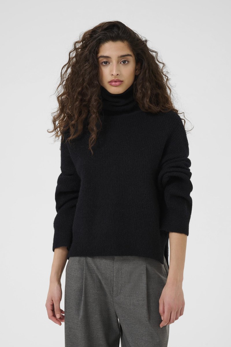 Black high-neck sweater with a textured finish, featuring dropped shoulders and ribbed cuffs. Paired with light gray trousers.