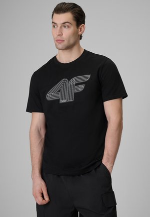 Mężczyzna w czarnej koszulce z białym stylizowanym logo "4F", stoi z jedną ręką w kieszeni, lekko patrząc w lewo.