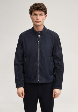 Giovane uomo con capelli castani ricci che indossa una giacca con zip blu scuro e pantaloni neri, in piedi davanti a uno sfondo chiaro e uniforme.