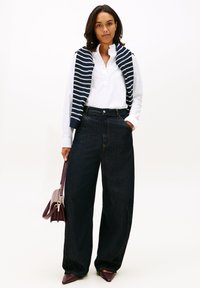 Žena v bílé blůze a širokých tmavých džínách, přes ramena přehozený pruhovaný svetr, drží tmavou koženou kabelku.