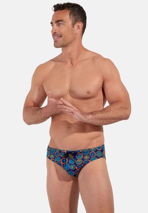 Muscular man smiling and looking left, wearing blue patterned swim briefs with orange circular designs, standing against a plain background.