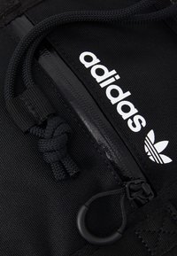 Närbild av svart Adidas-väska med dragkedja, flätade dragningssnören och vit Adidas-logotyp samt trefolk-emblem på texturerad tyg.