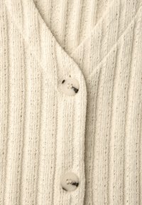 Krémově zbarvený pletený cardigan s vertikálním žebrováním a dvěma velkými lesklými knoflíky zapnutými uprostřed.