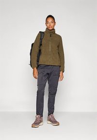 Naine seisab oliivirohelises fliisjakis, tumedates cargo-pükstes, pruunides matkingiides ja musta seljakotiga, neutraalse heleda tausta ees.