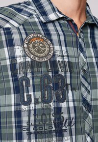 Blauwgroene geruite shirt met knopen, voorzien van een "Camp David C.63" patch en geborduurde outdoor-thema tekst op de borst.
