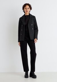Giacca nera doppiopetto con ampe rever, top nero testurizzato, pantaloni neri a vita alta e scarpe platform nere spesse.