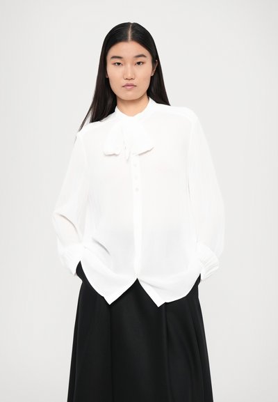Bluză albă cu fundă la guler, cu nasturi pe față și mâneci lungi, asortată cu o fustă neagră. Material transparent, croială lejeră.