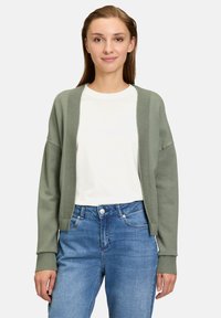 Vihreä neulecardigan, jossa on avoin etuosa, joustavat hihansuut ja rento istuvuus, pukeutuneena valkoisen t-paidan ja sinisten farkkujen päällä.