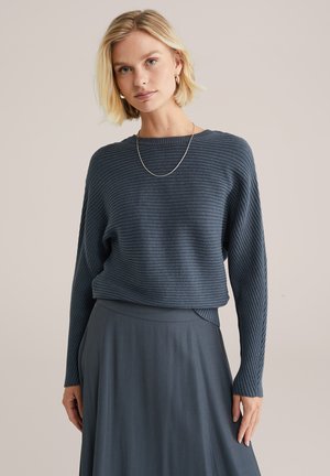 Pulover din tricot ribatat de culoare albastru închis, cu mâneci lungi, decolteu rotund și o croială ușor scurtă, asortat cu o fustă fluidă în aceeași nuanță.