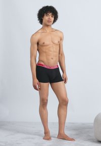 Young man with curly hair standing barefoot wearing black Tommy Hilfiger boxer briefs with red waistband.