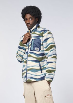 Man with curly hair wears a multicolor blue, green, and cream patterned fleece jacket with a navy chest pocket and beige cargo pants.