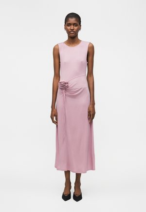 Naine seismas, kandes õlata heledat roosat midikleit kangast roosiga ja mustade teravate kontsakingadega valgel taustal.